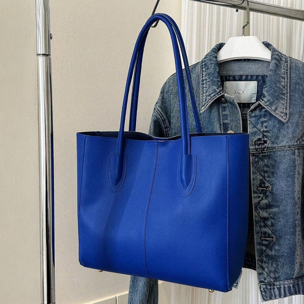 Blue minimalist structured tote bag with double handles, hanging on a rack beside a denim jacket.