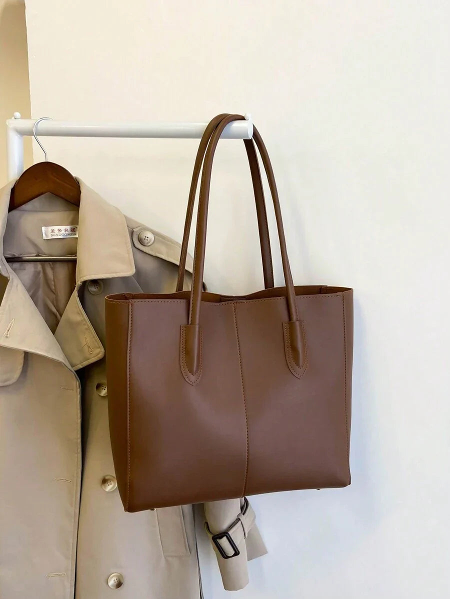 Minimalist brown PU leather shoulder tote bag with double handles hanging against beige coat on hanger
