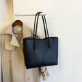 Black Carla Shoulder Tote bag hanging on a rack next to a beige trench coat, showcasing its sleek, minimalist design.