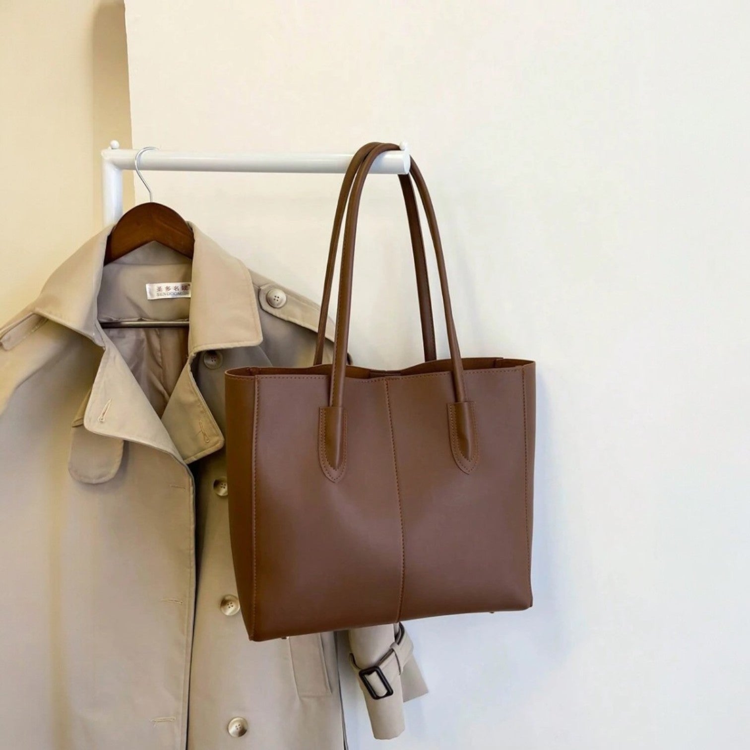 Brown Carla shoulder tote bag hanging on a rack next to a beige trench coat against a plain wall.