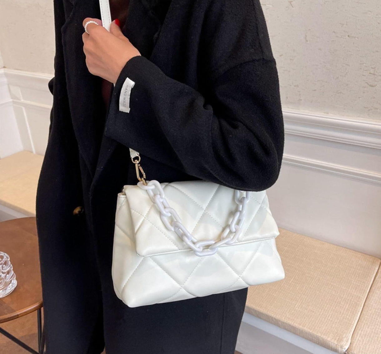 White quilted handbag with chunky chain detail, held by person wearing a black coat indoors