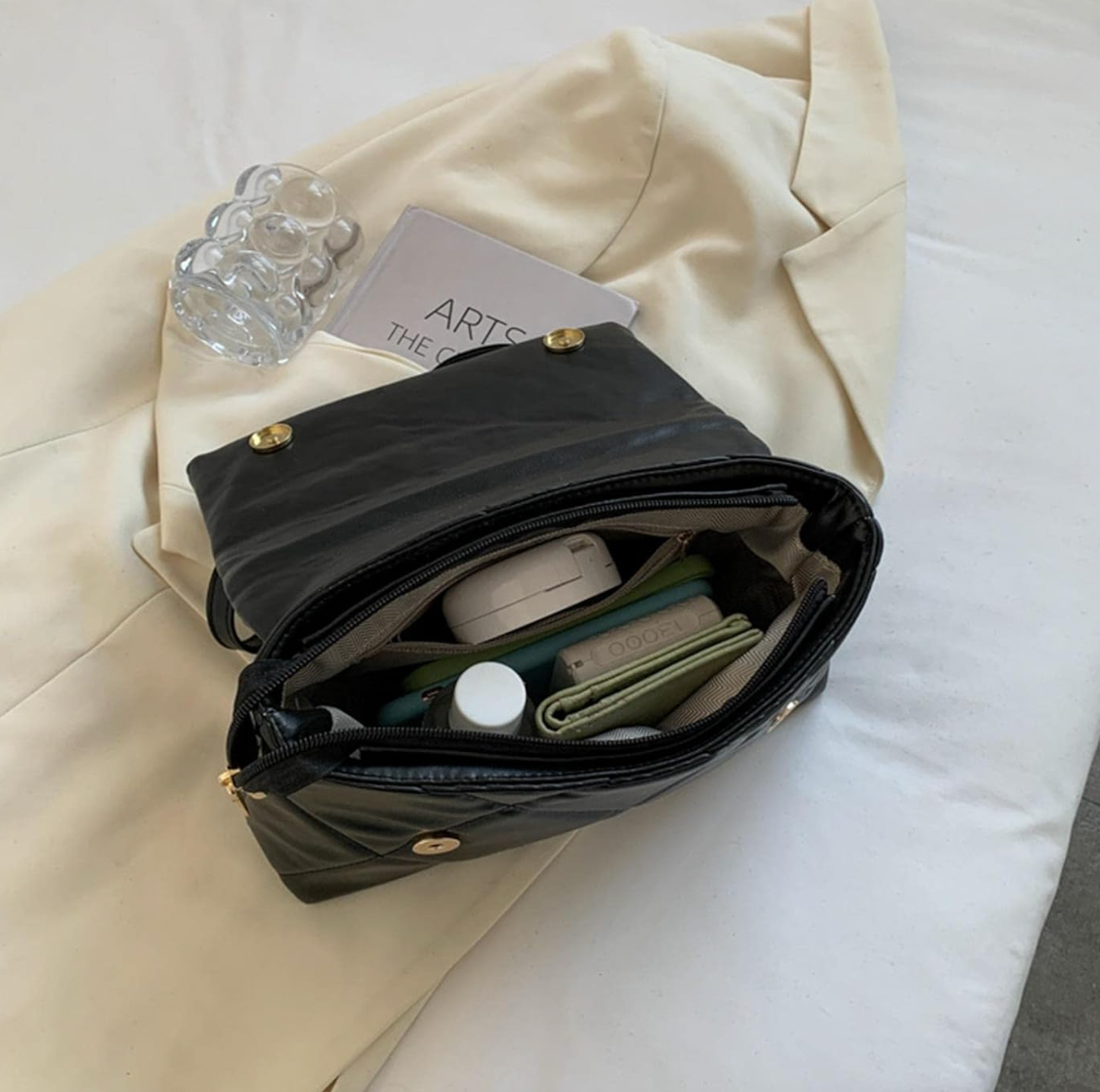 Open black quilted handbag with gold buttons showing organized interior items on a light beige jacket background