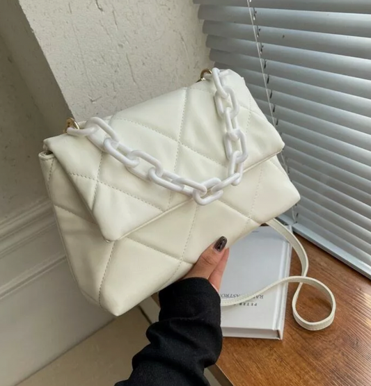 Quilted white handbag with chunky chain detail and shoulder strap held against a wall near window blinds