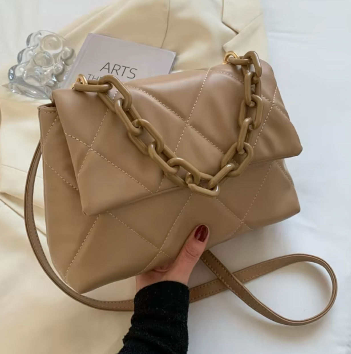 Beige quilted handbag with chunky chain detail and long strap, held over cream fabric background.